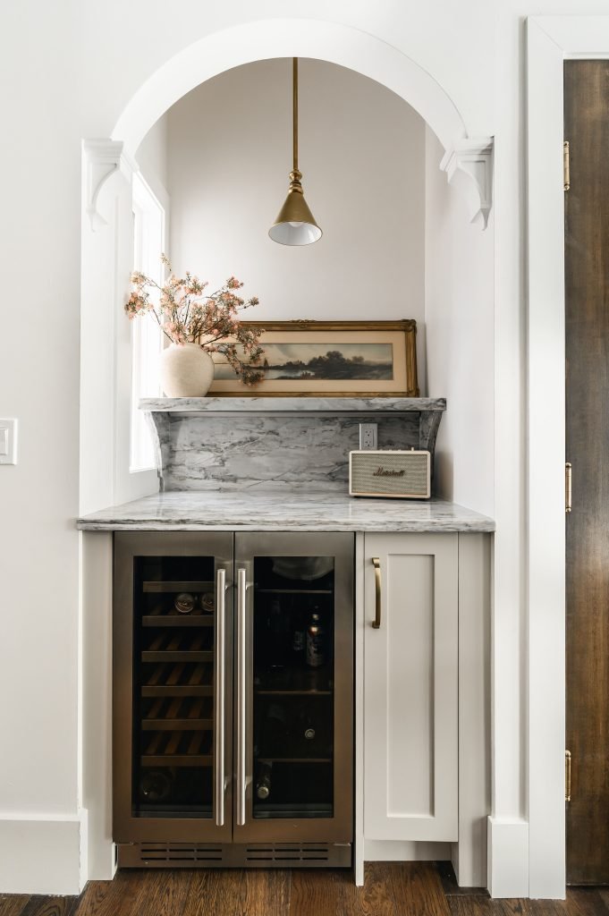 Built-in arched wine bar nook with a marble countertop, dual door wine fridge, vintage landscape painting, and a brass pendant light.