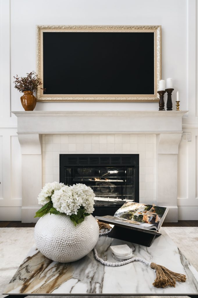 Bright fireplace surround with a gold-framed TV, flowers and candles on the mantle, and a marble coffee table in the foreground.