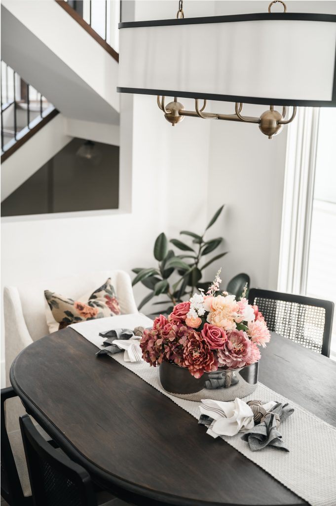 Dark oval dining table with a light runner, floral pink centerpiece in a metal vessel with oval light fixture above.