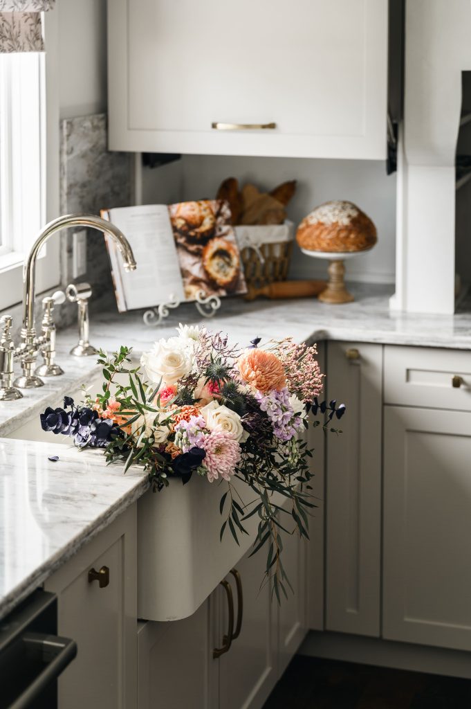 White apron farmhouse sink with a colourful wildflower arrangement in a kitchen with marble countertops.