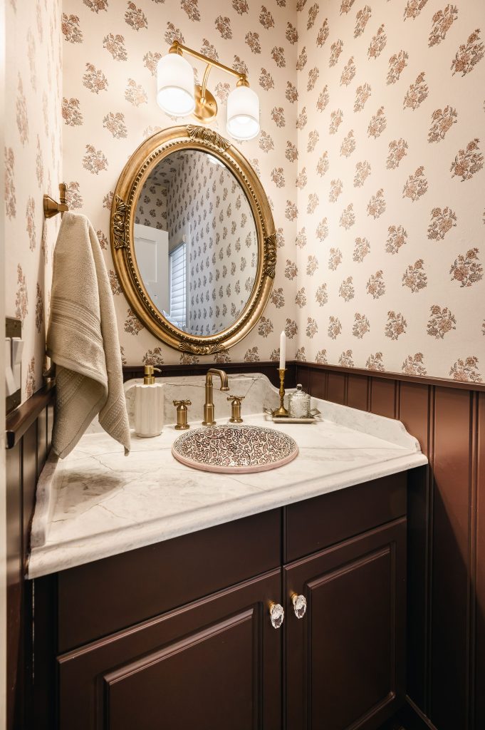 Victorian-inspired powder room with floral print wallpaper, dark wainscoting, and a gilded oval mirror above the vanity.