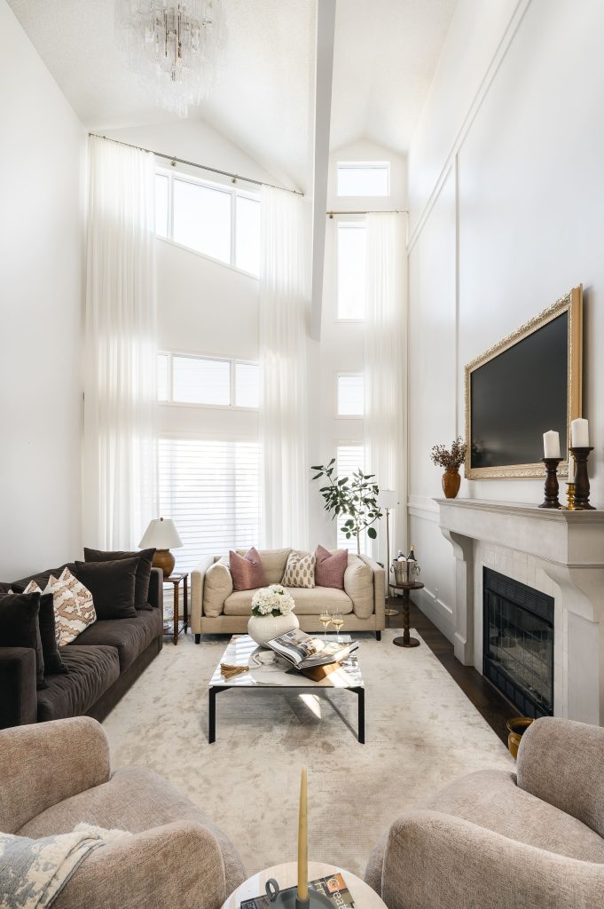 Grand double-height living room with a fireplace, gold-framed TV, sheer floor-length curtains, and a chandelier above.