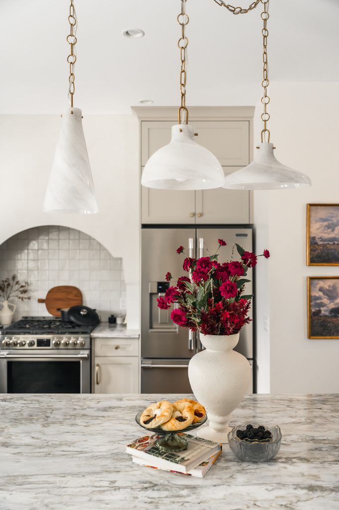 White glass pendant lights on brass chains hanging above a marble kitchen island with a white vase of bold red flowers.