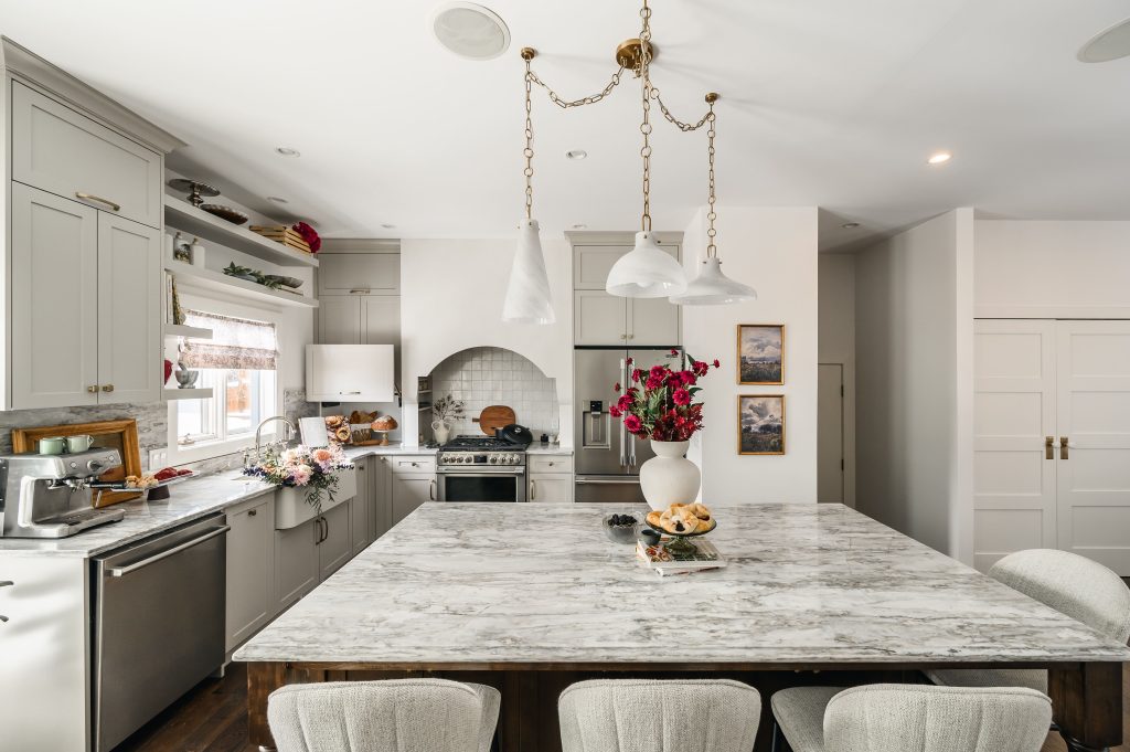 Large marble kitchen island with a dark wood base and cream bar stools beneath white pendants, with light cabinetry beyond.