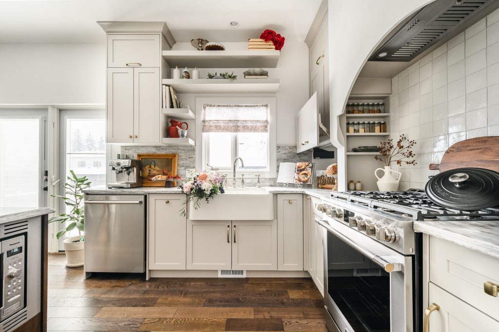 Bright kitchen corner with a white apron farmhouse sink, open shelving above, and shaker cabinets with marble countertops.