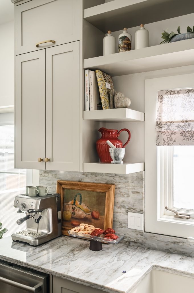 Open kitchen shelves styled with a red porcelain pitcher, books, and ceramics above an espresso machine on a marble counter.