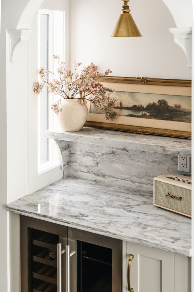 Marble-topped built-in bar counter with a vase of pink florals, a vintage landscape painting, and a brass cone pendant.