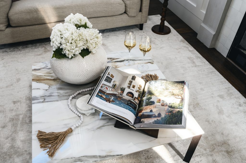 White marble coffee table styled with a white hydrangea vase, open design book, wine glasses, and beaded tassel in the living room.