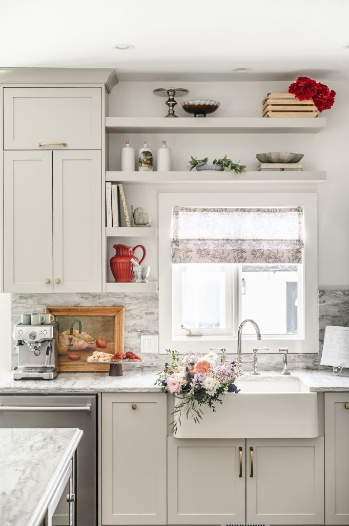 Kitchen sink wall with open shelves styled with ceramics, a red pitcher, and books, above a white apron farmhouse sink.