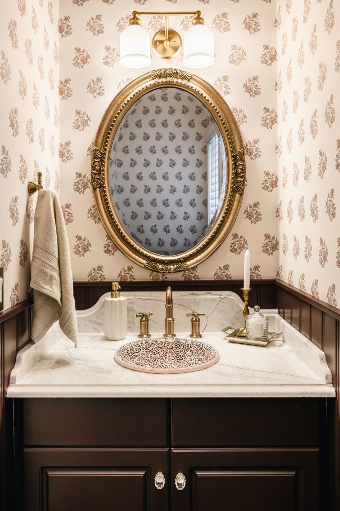 Full powder room vanity with a gilded oval mirror, floral wallpaper, dark wainscoting, and a sink on marble.