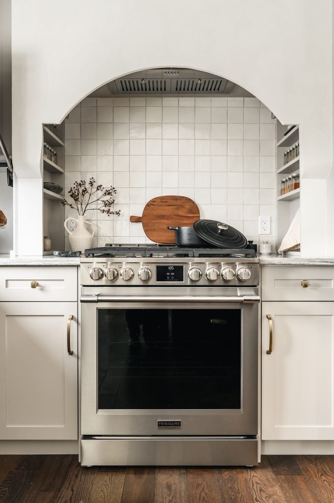 Close-up of an arched range hood with white square tile backsplash, wooden cutting board, and a cast iron Dutch oven.