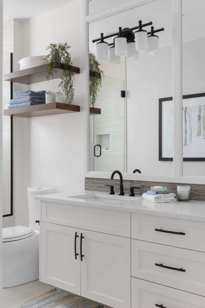 A white bathroom vanity with black hardware and a framed mirror sits beside open wood shelving holding folded towels and greenery. A framed piece of art and black vanity light complete the space.