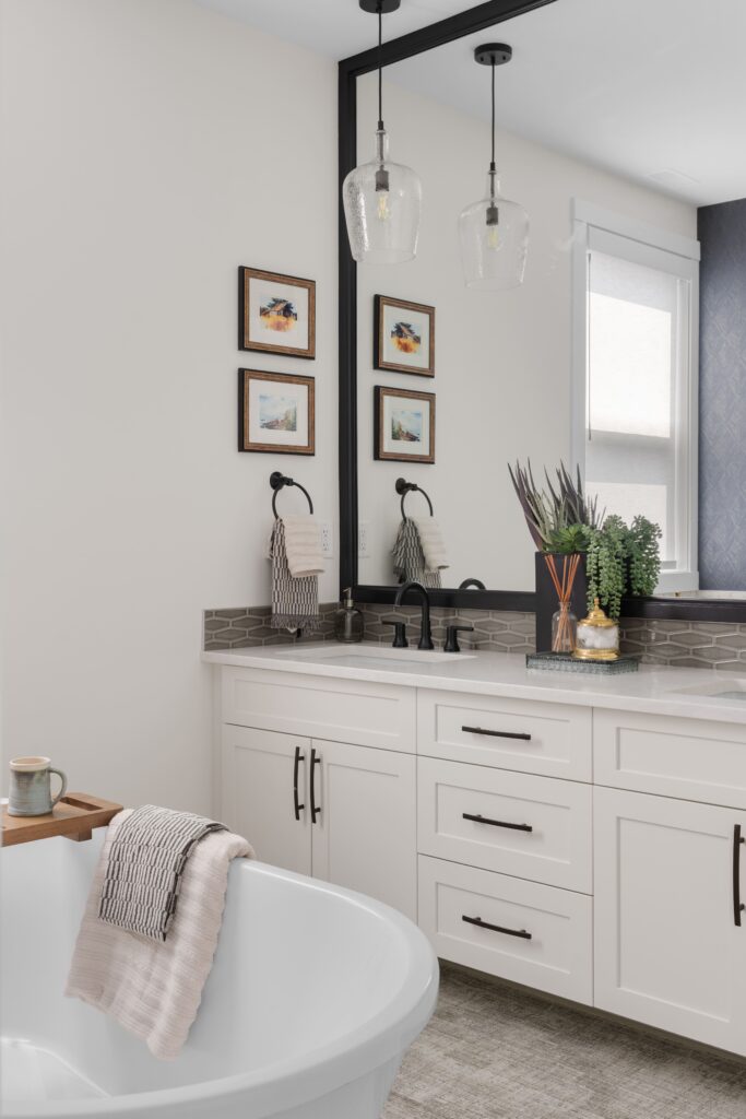 A white vanity with black hardware sits beneath a large mirror and a glass pendant light, styled with a stacked set of small framed landscape prints on the wall beside. A white freestanding tub and a coffee mug on a wood tray are visible in the foreground.
