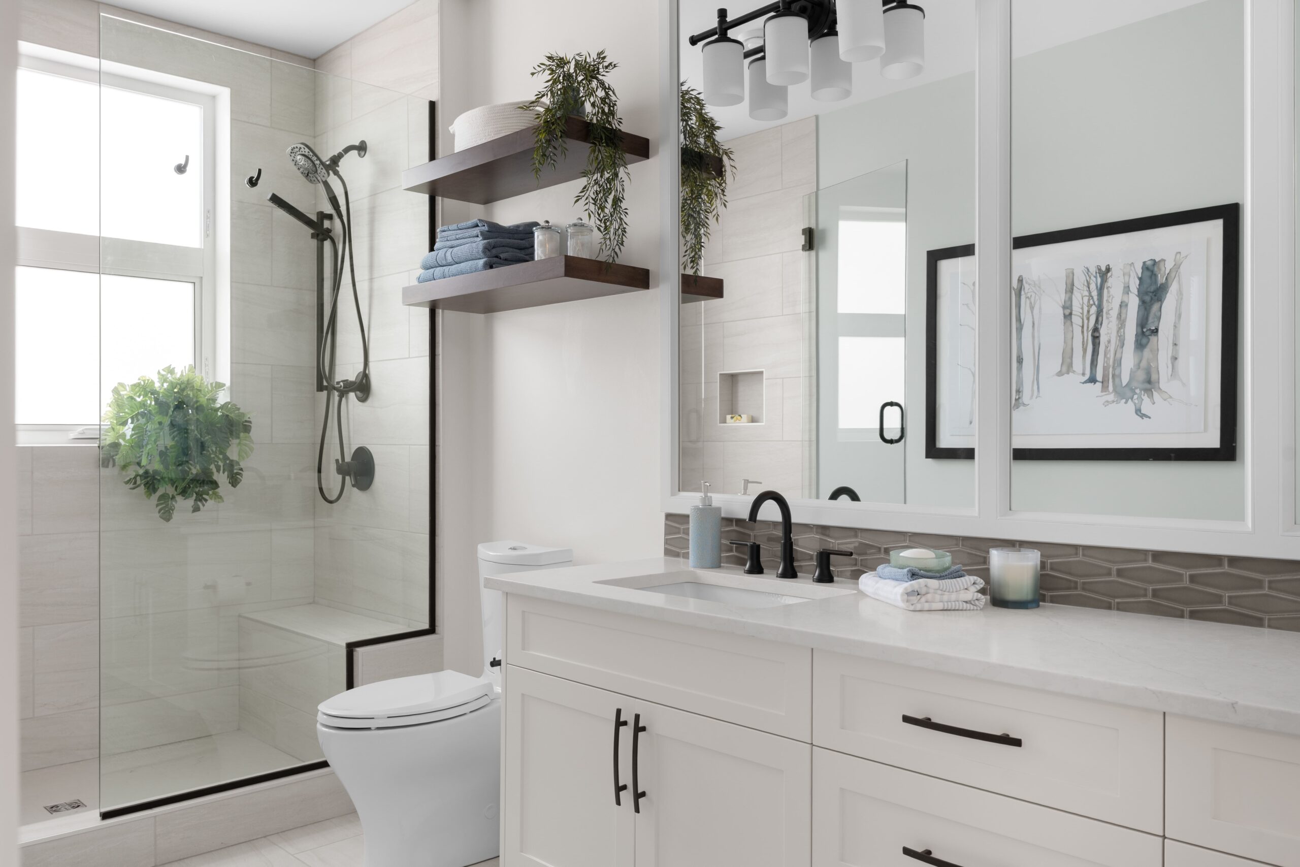 A bathroom features a glass-enclosed shower with a black rain showerhead and handheld sprayer, positioned beside a white vanity with a black faucet and undermount sink. Open wood shelving above holds towels and greenery, and a framed botanical print hangs on the opposite wall.