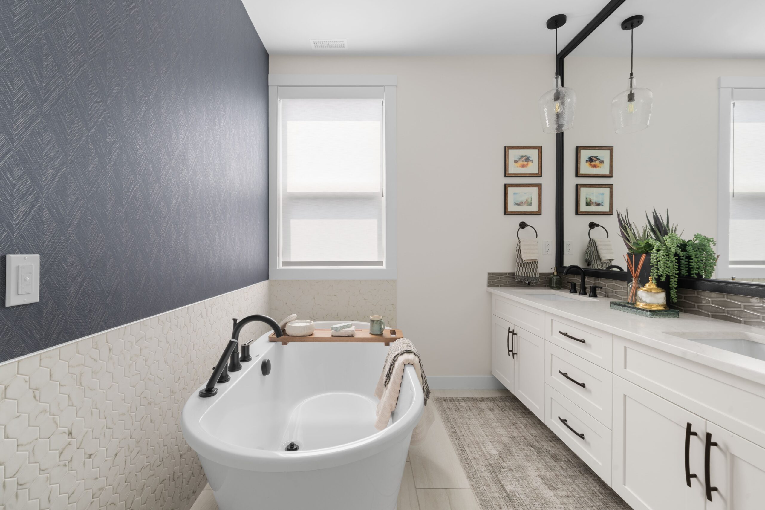 The ensuite bathroom features a white freestanding soaking tub with matte black fixtures beneath a dark textured wallpaper accent wall, positioned beside a white double vanity with black hardware. A glass pendant light hangs in front of the mirror, and small framed landscape prints decorate the wall.