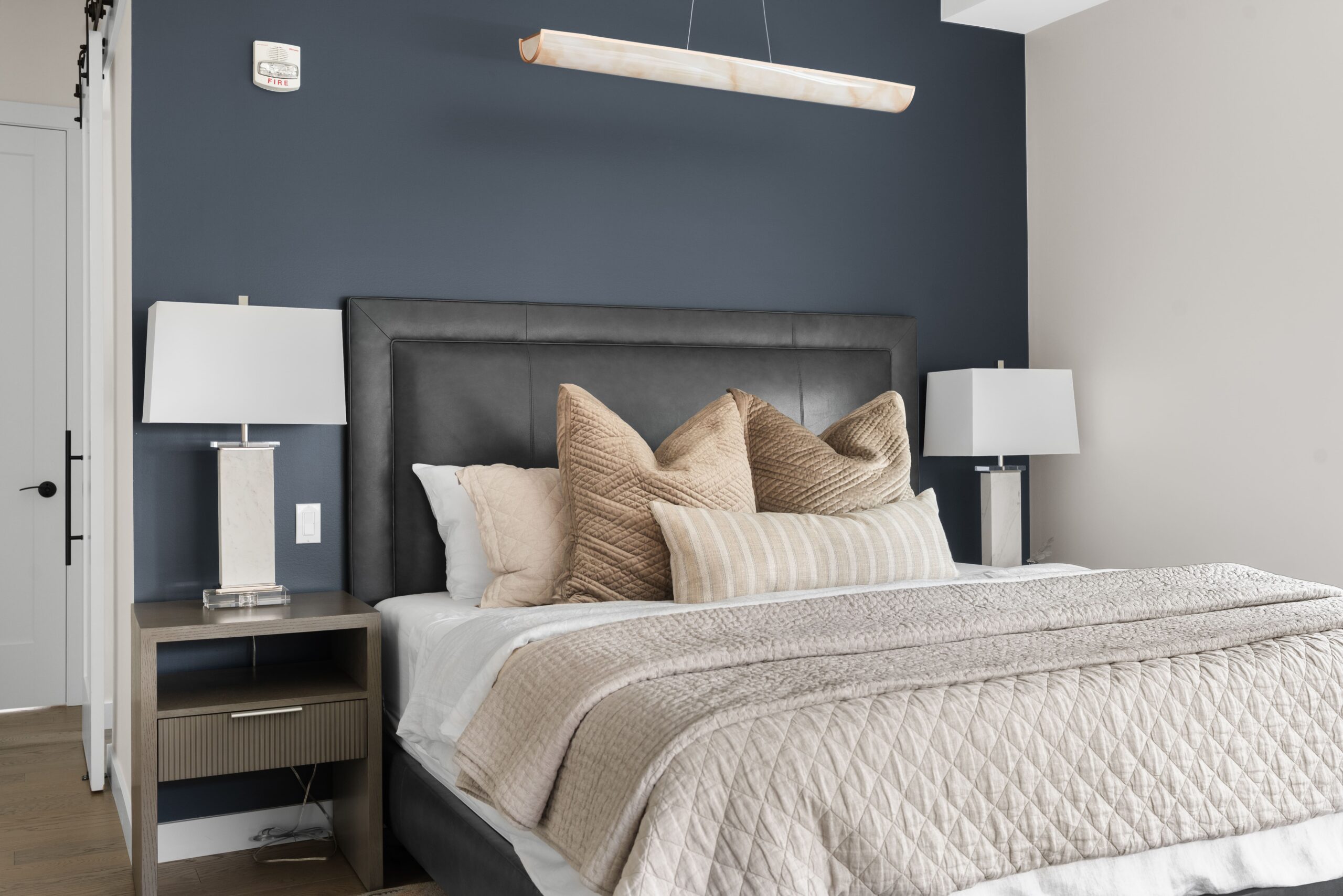 A bedroom features a gray upholstered headboard against a navy accent wall, dressed with layered neutral and taupe pillows and a quilted coverlet. Nightstands with white table lamps flank the bed beneath a linear alabaster pendant light.