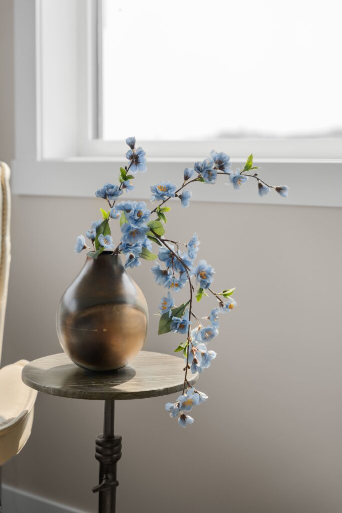 A bronze ceramic vase holds a branch of light blue faux flowering stems, displayed on a round wood pedestal side table in front of a window. A neutral reclining chair is partially visible at the edge of the frame.