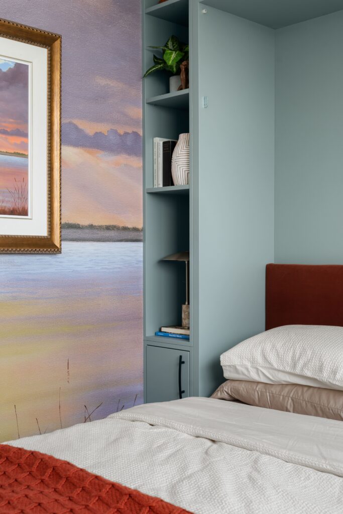 Built-in shelving holds a small plant, a vase, and books beside a bed with a rust-colored upholstered headboard and layered white and taupe pillows. A framed sunset landscape painting and matching wallpaper mural are visible at left.