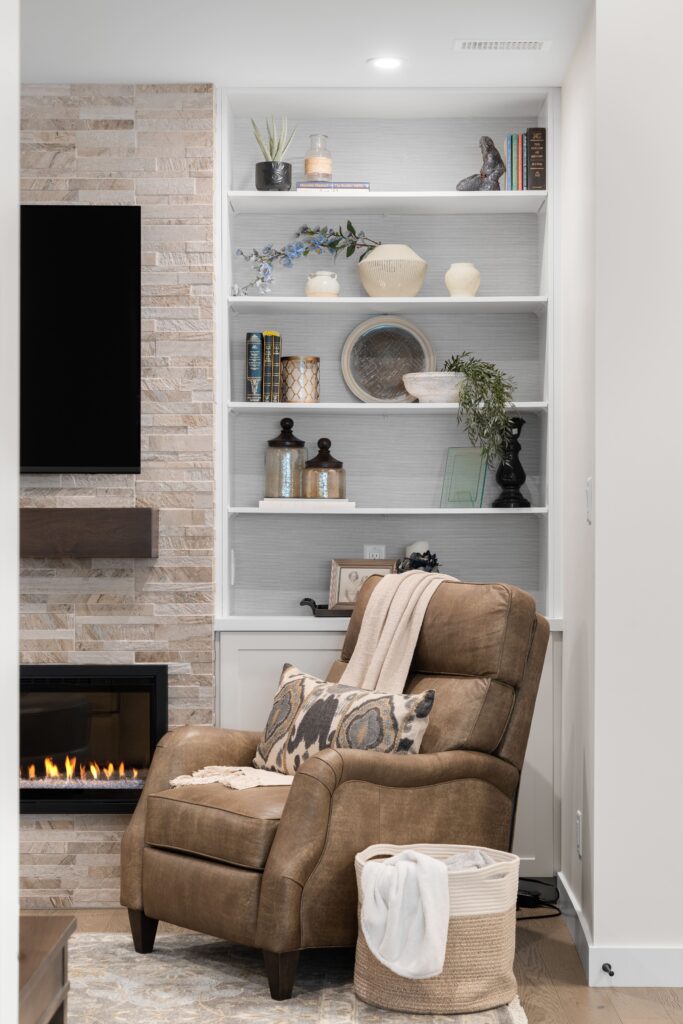 White built-in wallpapered shelving displays books, ceramic vases, and decorative objects beside a stacked stone fireplace with a mounted television above. A brown leather recliner with a patterned throw pillow and a woven storage basket sits in front of the shelving.