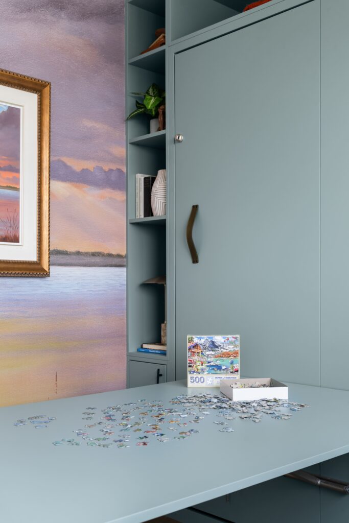 A close-up of built-in cabinetry with a leather murphy bed handle and open shelving holding a vase and books, positioned above a fold-down desk surface with a puzzle in progress. A framed sunset landscape painting and custom wallpaper mural are visible in the background.