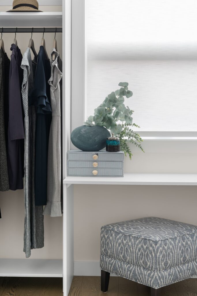 A built-in closet vanity nook holds a grey ceramic vase with faux eucalyptus and a small candle, sitting on top of a patterned storage box on a white vanity desk. A patterned upholstered ottoman sits below, with a hanging rod of clothing visible at left.