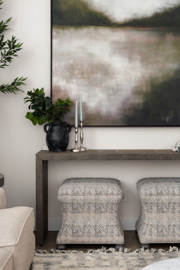 A dark console table sits below framed artwork, styled with a black textured vase holding greenery and a pair of silver candlesticks. Two patterned upholstered ottomans sit beneath the console.