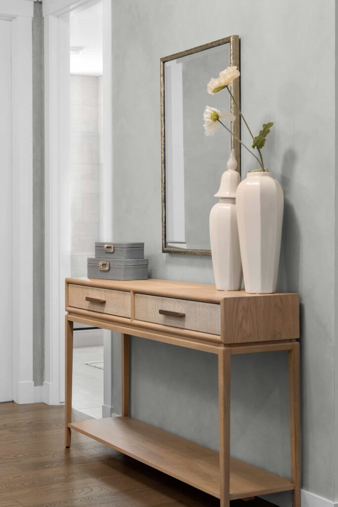 A light wood console table with two drawers holds a pair of white ceramic vases with faux white flowers, gray storage boxes, and a wall mirror above. The setup sits against a soft gray wall near a hallway opening.