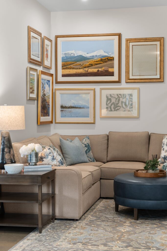 A gallery wall of framed landscape and botanical art hangs above a taupe sectional sofa styled with blue and floral throw pillows, set beside a navy leather round ottoman. A dark wood side table with a table lamp and faux white flowers completes the seating area.