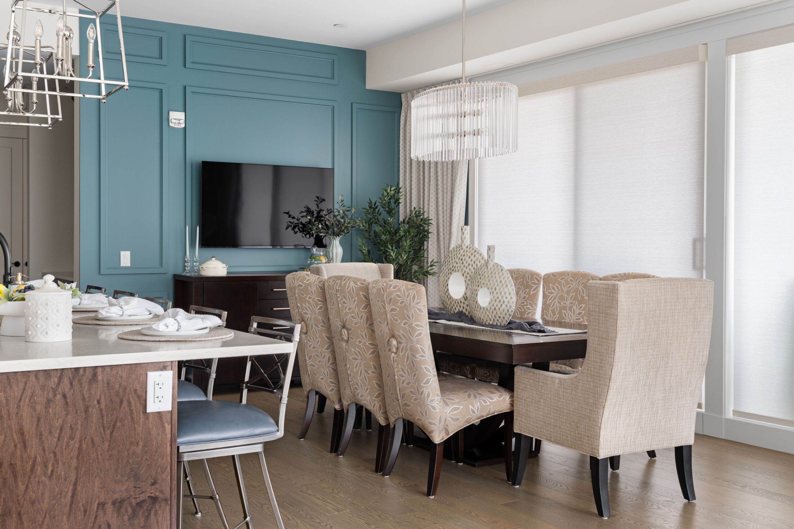 A dining area features a long table with upholstered floral-patterned chairs beneath the chandelier, set beside a kitchen island with a chrome lantern-style pendant light. A teal paneled accent wall with a mounted television and dark wood console anchors the far side of the room.