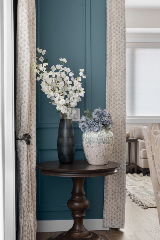 A teal paneled corner holds a dark wood table styled with a black vase of white flowering branches and a mosaic vase of blue hydrangea. Floor to ceiling curtain panels frame the corner with a dark metal curtain tie back.