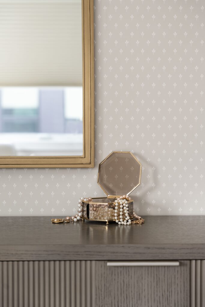 A glass jewelry box with pearl necklaces sits open on a wood dresser beneath a gold-framed mirror, set against a small floral dot wallpaper pattern. The vignette highlights a bedroom vanity styling area.