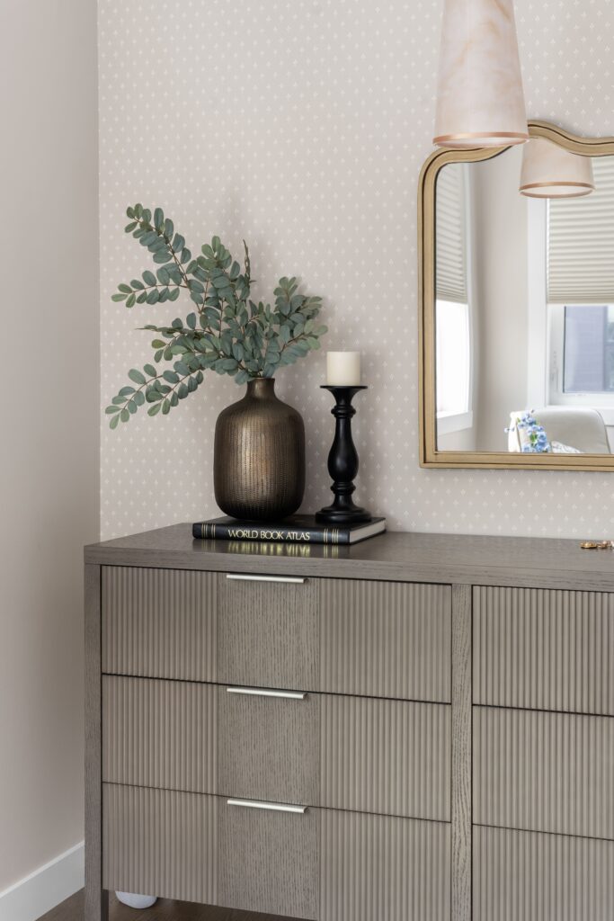 A close-up of a fluted wood dresser displays a dark textured vase with faux eucalyptus, a black candlestick with a pillar candle, and a book, set against a cream dotted wallpaper. A gold-framed mirror reflects a window in the background.