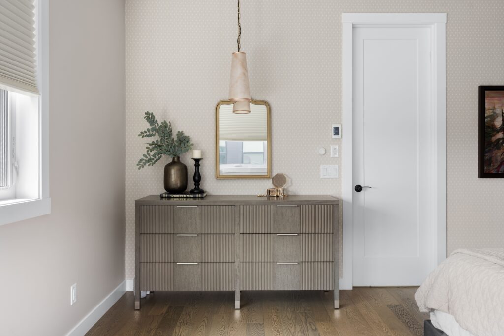 A fluted wood dresser with metal drawer pulls holds a dark ceramic vase with faux greenery, a candle, and a small decorative box, set beneath a gold-framed mirror. An alabaster pendant light fixture hangs above the dresser.