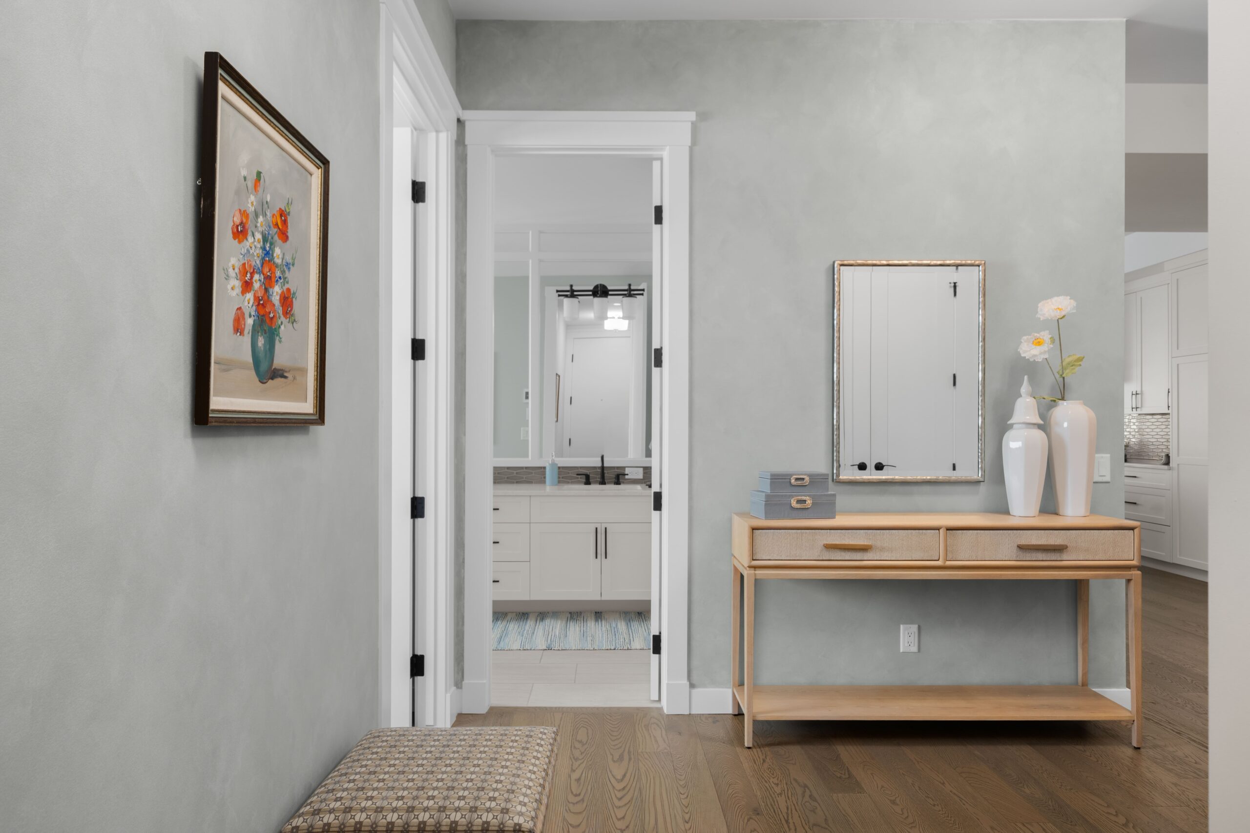 A light wood console table with a mirror in the hallway entry. A doorway opens to a bathroom with white cabinetry visible beyond, and a patterned ottoman sits in the foreground.