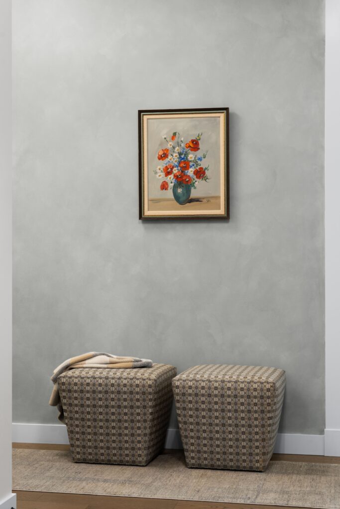 A framed floral still-life painting hangs on a soft gray wall above two patterned upholstered ottomans, one draped with a folded throw blanket. The vignette sits beside a woven area rug in the entryway.