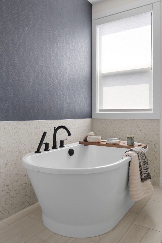 A white freestanding soaking tub with a matte black faucet and handheld sprayer sits below a window, framed by a dark textured wallpaper accent wall and a mosaic tile surround. A wood tray across the tub holds soap, a mug, and a candle.