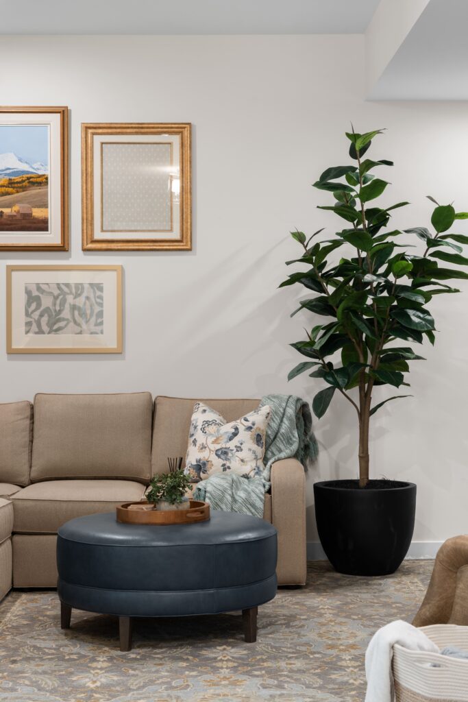 A portait view shows framed landscape and botanical art above a taupe sectional sofa with a floral throw pillow and a green knit throw, beside a tall faux fig tree in a black planter. A navy leather round ottoman with a wood tray sits in front of the sofa.