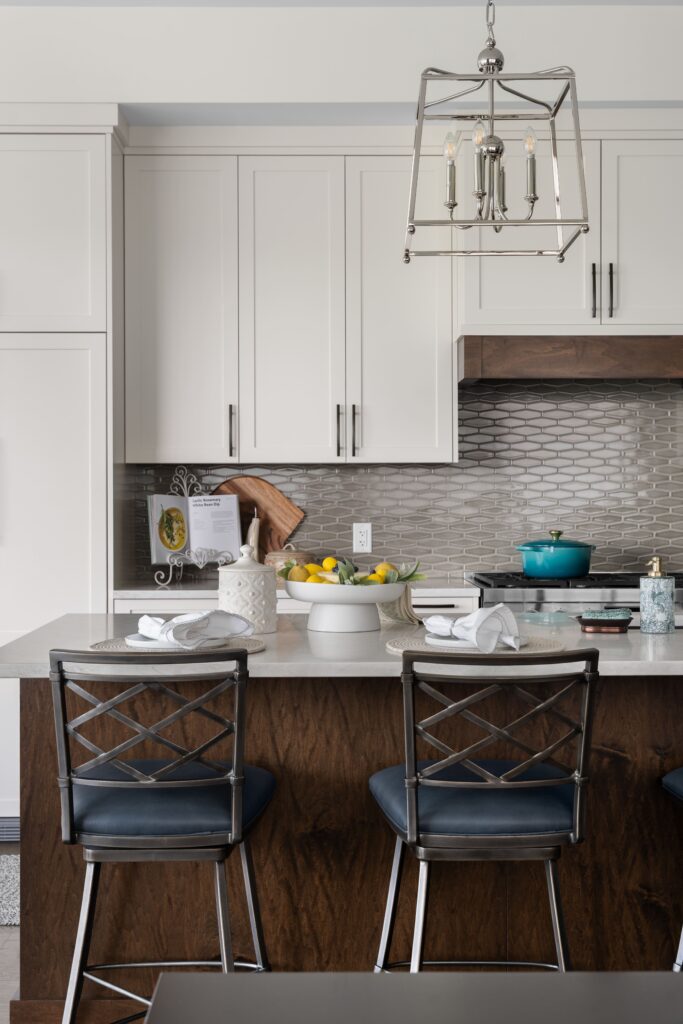 A white pedestal bowl of cheesecake and lemons sits on a light stone kitchen island counter near a range with a teal enameled pot, set beneath a lantern-style pendant light. White shaker cabinetry and a tiled backsplash frame the cooking area.