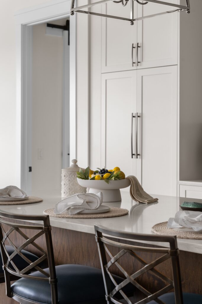 A white pedestal bowl filled with cheesecake, lemons, and bluberries sits beside a small ceramic jar on a light stone kitchen island counter, styled with woven placemats and folded napkins. White paneled cabinetry and a doorway are visible in the background.
