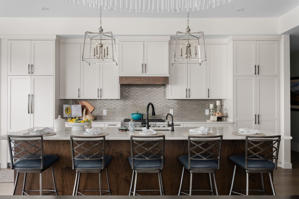 A kitchen features a large island with navy leather barstools, illuminated by two matching lantern-style pendant lights above a light stone countertop. White shaker cabinetry and a tile backsplash complete the space.