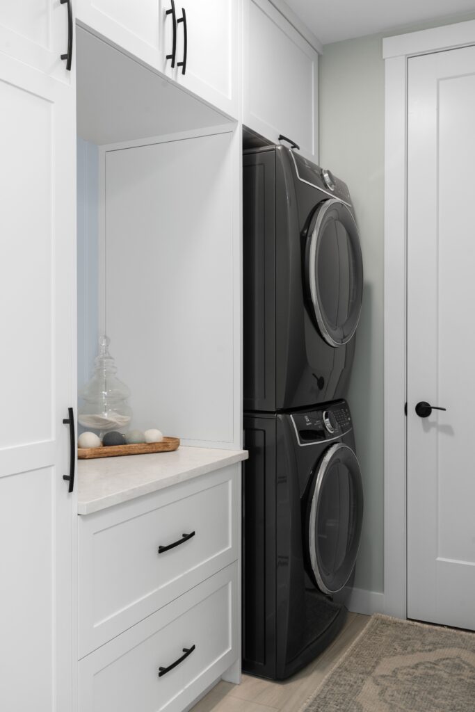 A laundry room houses a stacked washer and dryer beside white cabinetry with a stone countertop, a glass jar, and a wood tray. Black door hardware and open shelving complete the space, with a paneled door leading to an adjacent room.
