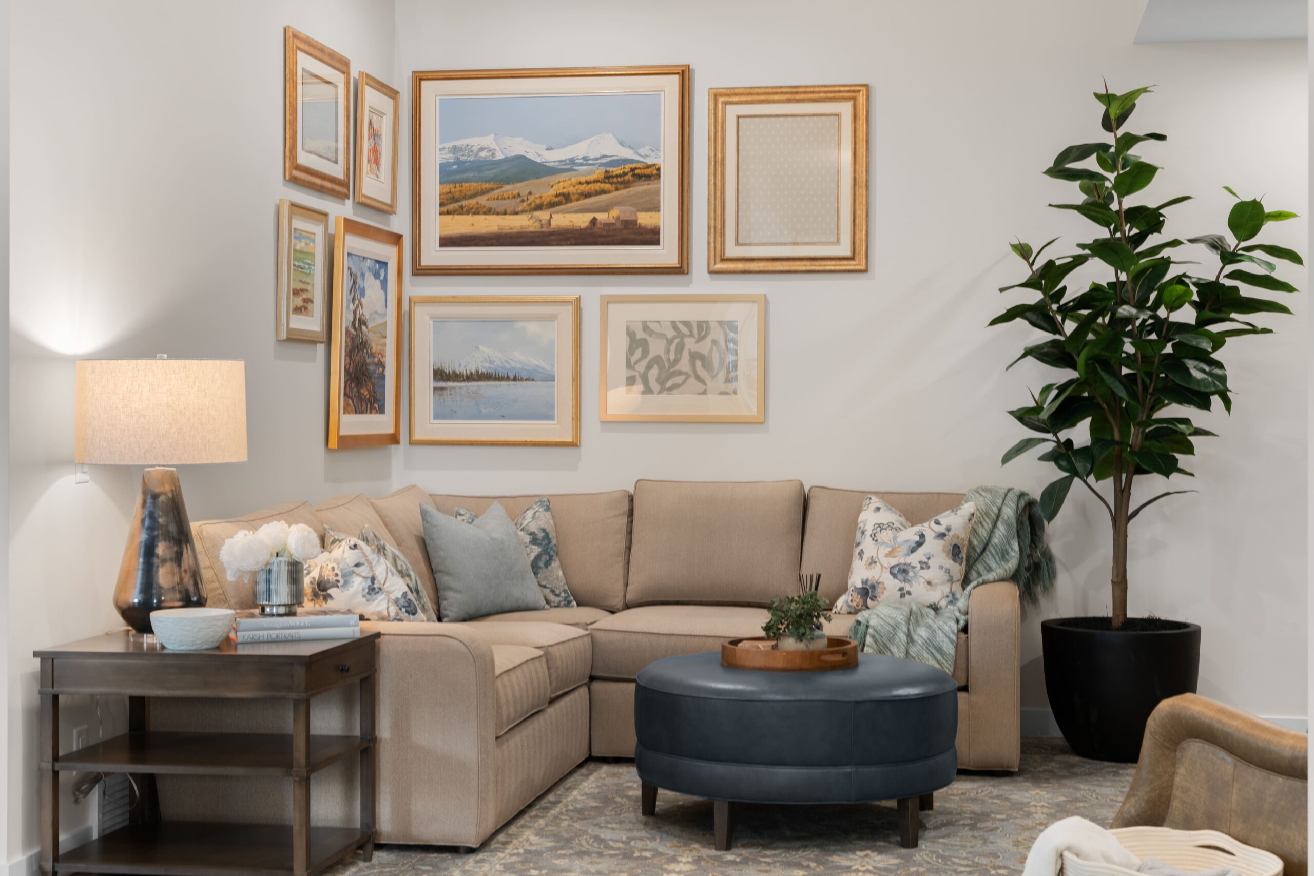 The living room features a gallery wall of framed landscape and botanical prints above a taupe sectional sofa, paired with a navy leather round ottoman and a tall faux fig tree in a black planter. A dark wood side table with a lamp and white flowers sits at the edge of the seating area.