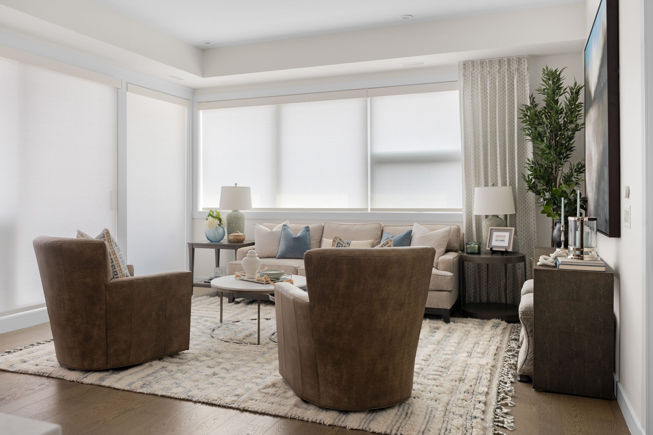 The living room seating area includes two brown leather chairs facing a cream sofa with blue and neutral throw pillows, arranged around a round coffee table on a woven area rug. Sheer window shades filter natural light along the back wall.