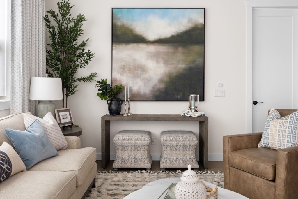 A living room features a framed landscape artwork above a dark wood console table styled with a black vase, greenery, and candlesticks. A cream sofa with blue pillows and a brown leather chair complete the seating area, arranged on a woven area rug.