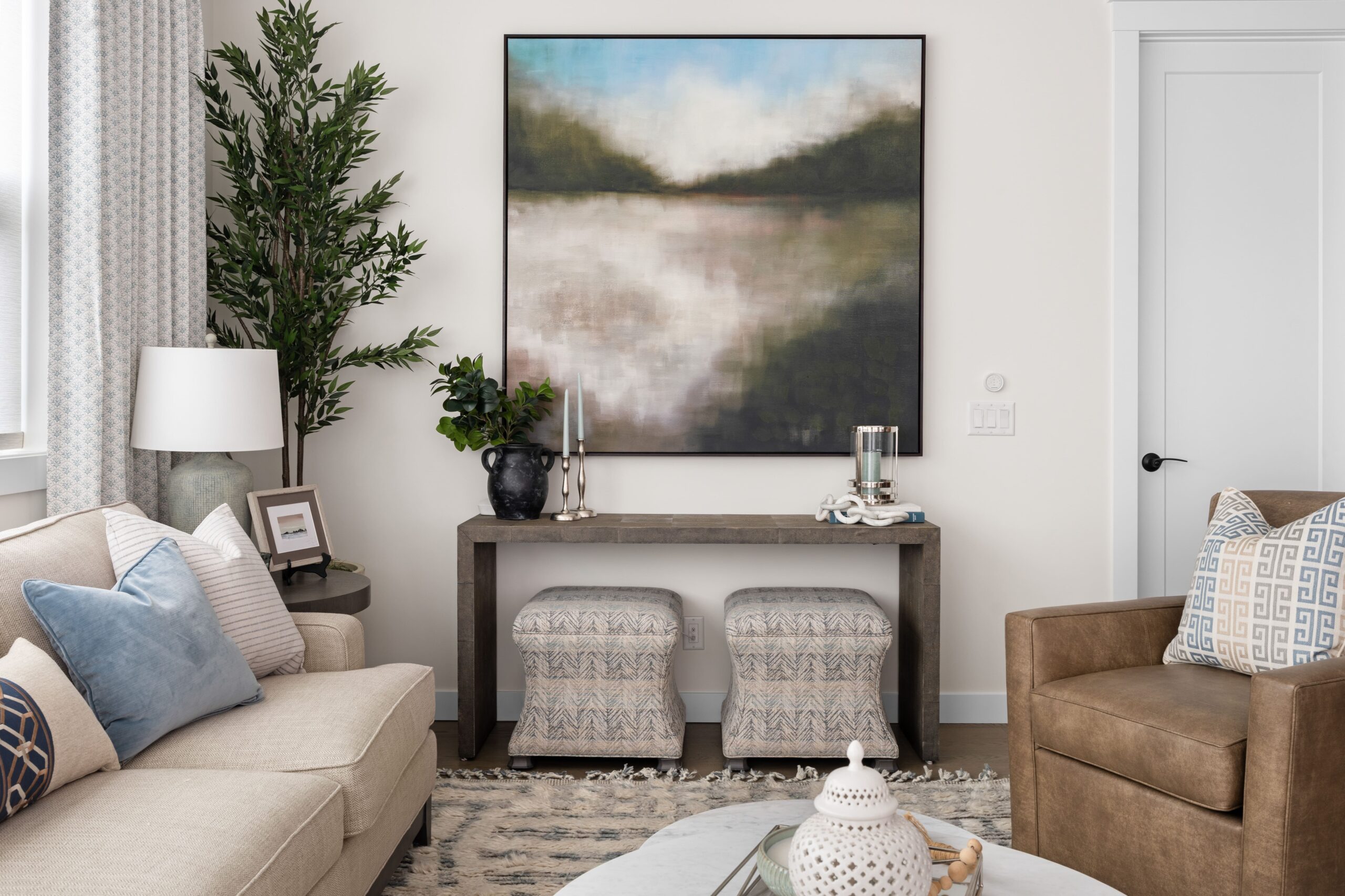 A living room features a framed landscape artwork above a dark wood console table styled with a black vase, greenery, and candlesticks. A cream sofa with blue pillows and a brown leather chair complete the seating area, arranged on a woven area rug.