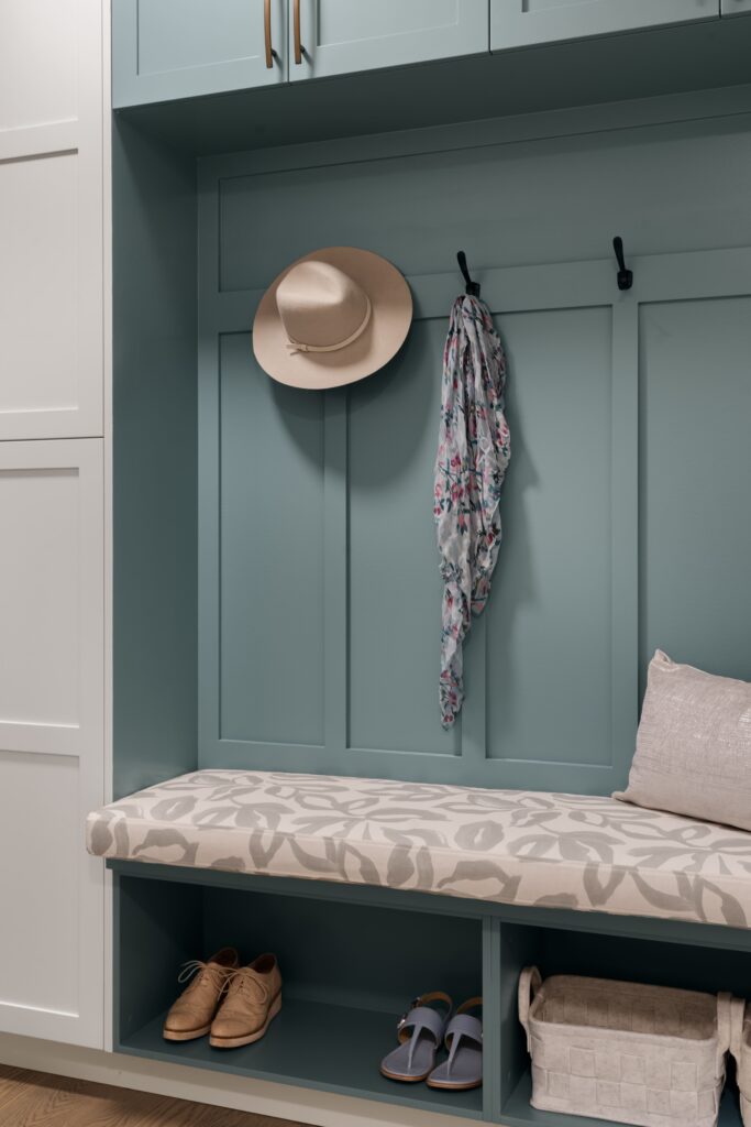 A tan hat and a floral-print scarf hang on black wall hooks above an upholstered mudroom bench in a neutral botanical-print fabric, set against teal paneled cabinetry.