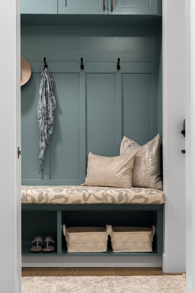 A teal-paneled mudroom nook features an upholstered bench cushion in a neutral botanical print, topped with a taupe pillow and a metallic patterned pillow. Woven storage baskets and a pair of sandals sit in the open cubby below.