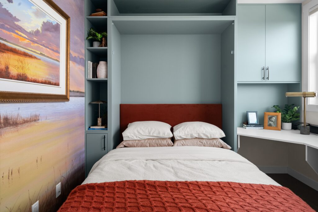 A bedroom features a murphy bed with a rust upholstered headboard flanked by built-in shelving and overhead cabinetry, with a corner desk nook holding a framed photo and a small potted plant. A sunset marsh custom wallpaper mural covers the wall to the left of the bed.