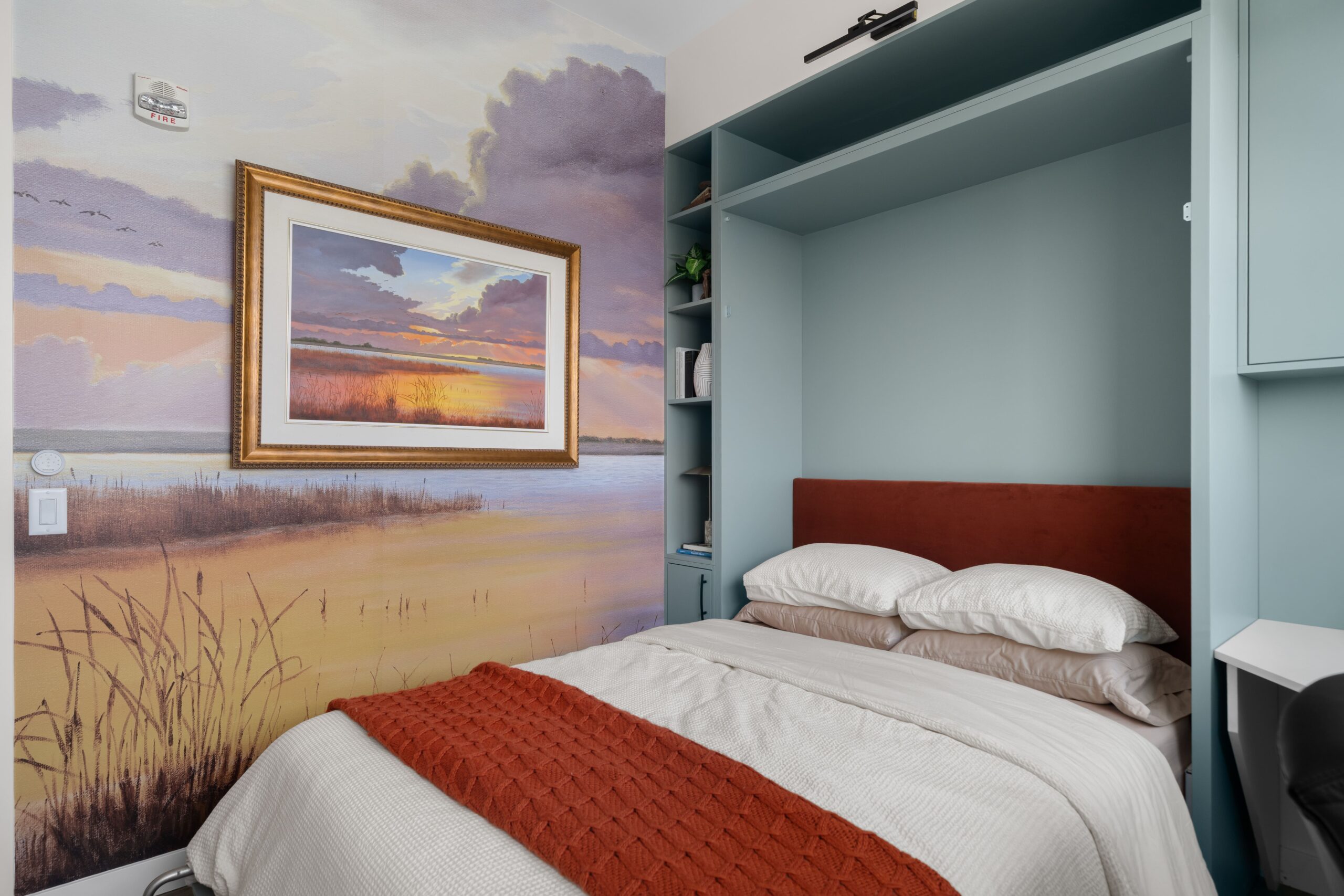 A framed sunset landscape artwork hangs beside a built-in murphy bed with a rust-colored upholstered headboard and light bedding topped with a rust knit throw. A custom wallpaper mural depicting a marsh at sunset extends across the adjacent wall.