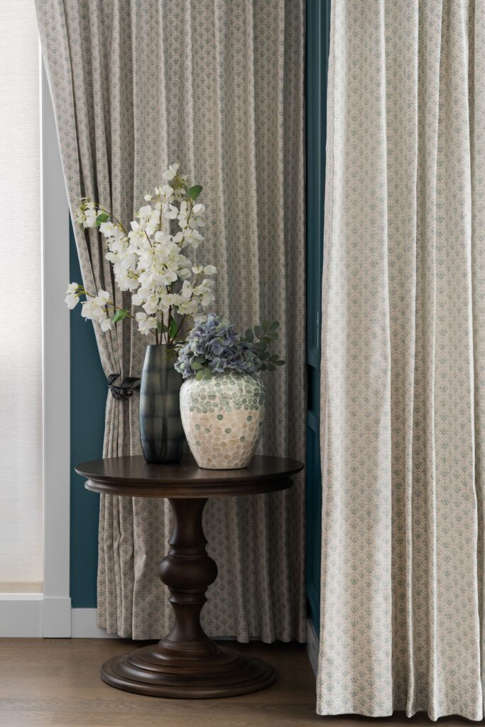 A dark wood pedestal table holds a black textured vase with white branch stems and a mosaic-patterned vase with blue hydrangea, set against a teal paneled wall. Two curtain panels with a small teal floral print frames the vignette.